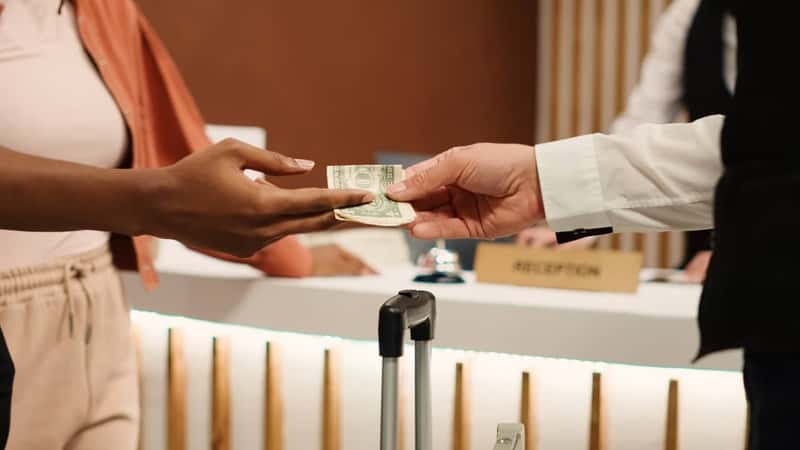 Close up of thankful hotel guest tipping helpful bellboy after receiving great customer service. African american woman handing dollar banknote to porter employee for helping with baggage