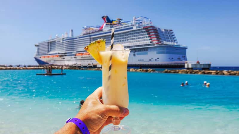 Close-up view of hand holding glass of alcoholic cocktail on Caribbean beach with cruise ship in background.