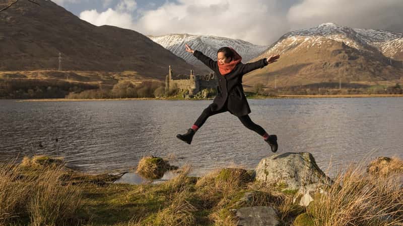 A girl wearing black clothes and a scarf from a rock on a lakeside with the Kilchurn Castle across the Loch Awe in the distance in the Scottish Highlands, UK, with snowy mountains on the background.
