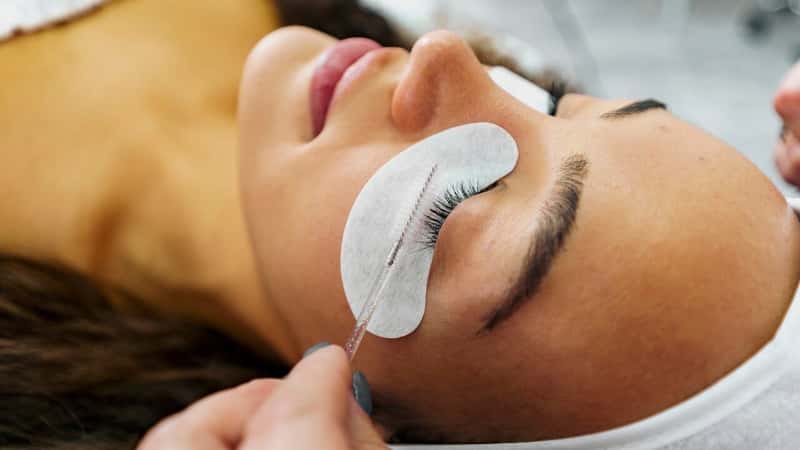 Attractive young woman getting her lashes done at the spa, laying on a massage bed. Beauticians hands doing clients eyelashes
