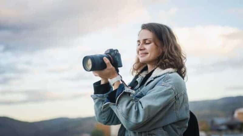 Young cheerful woman traveler in a denim jacket with a backpack holds a camera in her hands while standing on the shore of the mountain lake