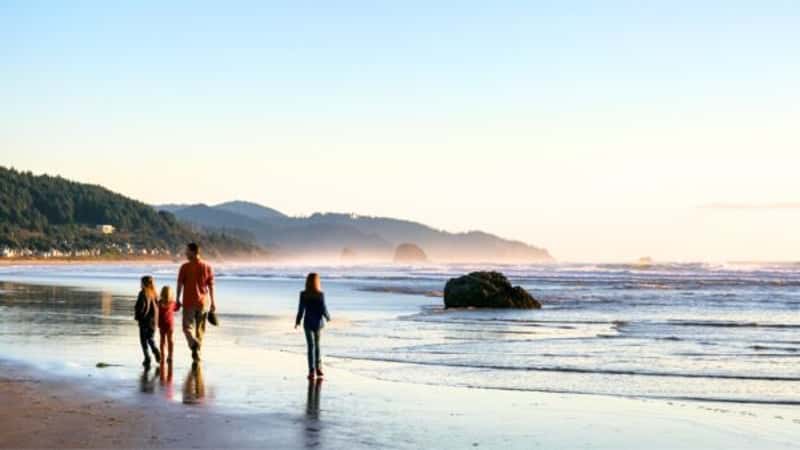 Cannon Beach, OR, showcases a family leisurely strolling along the water's edge during sunset. The scene captures the golden hues of the setting sun reflecting on the ocean waves.