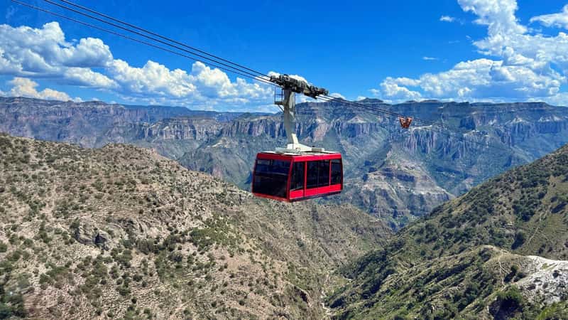 A sunlit panoramic view of Copper Canyon, with a cable car full of tourists capturing moments against the sprawling natural vista, under a nearly clear sky, with no identifiable traces of individuals