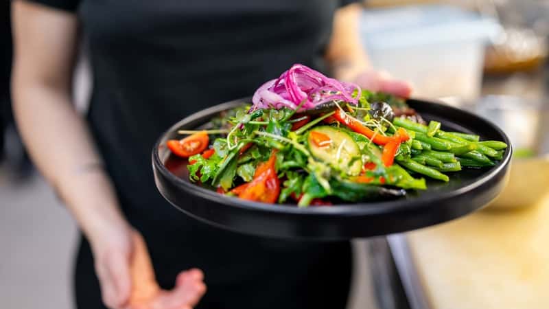 An image depicting an individual holding a vibrant salad plate. The plate contains fresh green beans, red tomatoes, and purple onions.