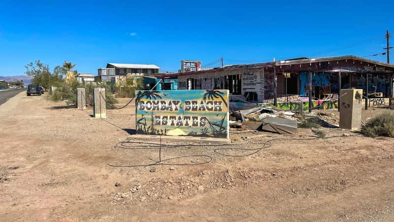 A photograph depicting buildings at Bombay Beach, California, featuring artistic designs. The image includes a view of the Bombay Beach Estates sign. The buildings exhibit creative elements, contributing to an artistic ambiance within the area.