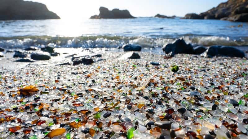 Millions of small pieces of old round-shaped glass cover the beach at Fort Bragg, California. The glass pieces create a colorful mosaic of greens, blues, whites, and browns. Ocean waves crash against the shore, with rocks scattered in the background.