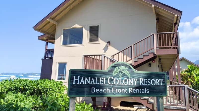 Kauai's Hanalei Colony Resort offers beach front rentals in a two-story building with a triangular roof. The structure is beige with a dark brown roof. In the lower foreground, a sign displays the hotel's name, "Hanalei Colony Resort - Beach Front Rentals."