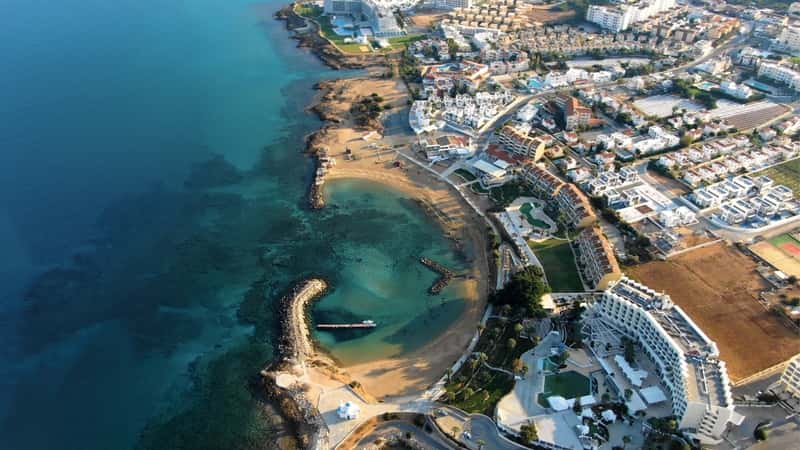 A drone-captured panorama showcasing the coastline of Larnaca, Cyprus. The image depicts a stretch of coastline featuring luxury hotels, villas, bays, and ports with stylish yachts. Sandy and rocky beaches line the calm sea with clear blue water, contributing to the overall scenic view. 