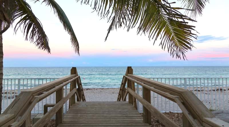 Vero Beach is a charming city located on the east coast of Florida. Close-up of the entrance to the beach at sunset with wooden steps and a beautiful fenced promenade with a white fence. The sand is white, the water is crystal blue, and the sky is iridescent shades of blue, white, and pink.