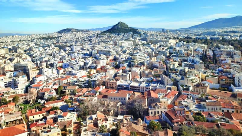 A panoramic view capturing Athens, Greece, with Mount Lycabettus prominently visible in the horizon. The lower section of the image features colorful houses representing the Anafiotika neighborhood, known for its picturesque charm.