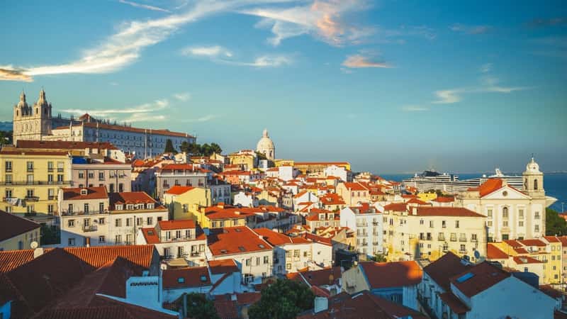 A photograph capturing the skyline of Alfama, the oldest neighborhood in Lisbon. The image showcases the architectural features of the district, characterized by a mix of historic buildings and narrow streets. The red-tiled roofs are a prominent element in the skyline, reflecting the traditional aesthetic of the area.
