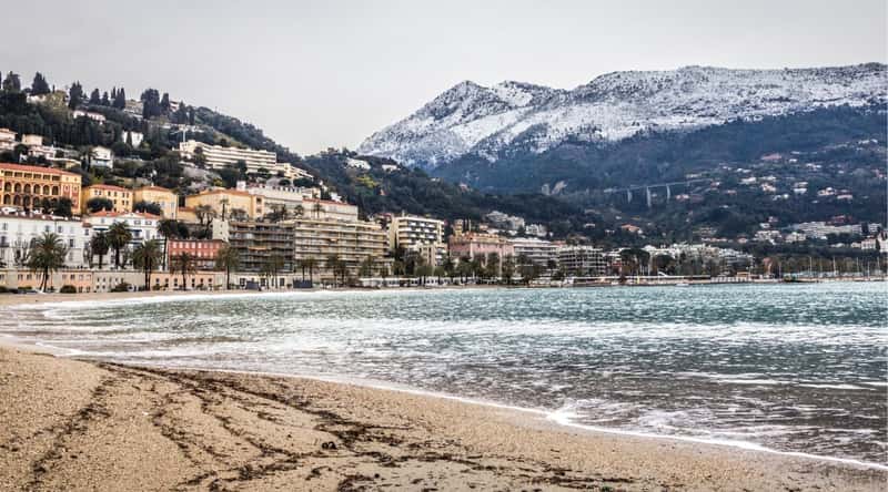 A photograph depicting the beach in Menton, France, set against the city backdrop. The image showcases the sandy shore and coastline with minimal foreground activity. In the background, the cityscape of Menton is visible.