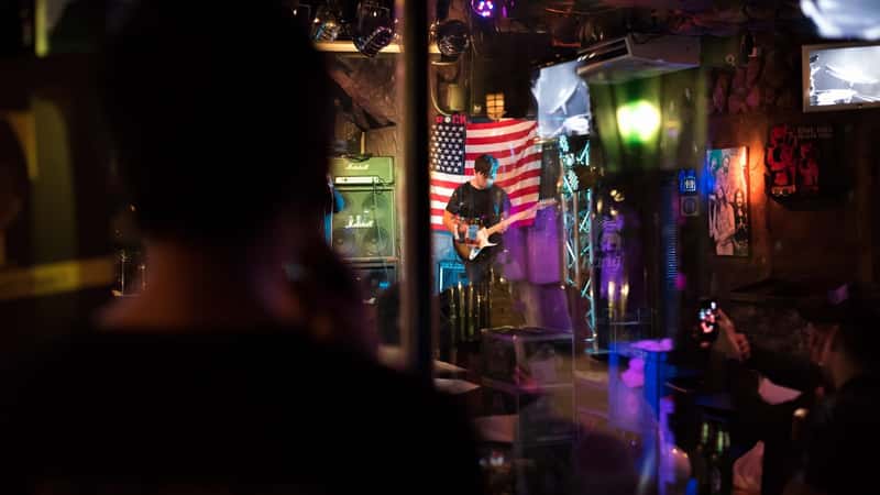 An image depicting a guitarist on stage in a small club setting. The musician is playing the guitar, with a backdrop featuring a United States flag on the wall behind him. The audience is comprised of individuals who are watching and listening to the performance. The setting suggests an intimate club environment, with the focus on the guitarist and the patriotic backdrop.