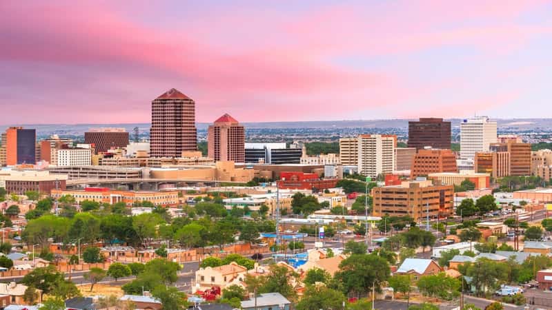 Albuquerque, New Mexico, USA, downtown cityscape at twilight features low buildings in the foreground surrounded by greenery and trees. In the background, the city rises with high buildings, illuminated against the twilight sky.