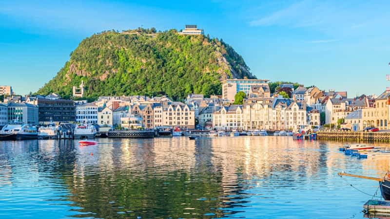 The image captures a view of Alesund, Norway, from the port during sunset, located in More and Romsdal County. The town is situated on the seashore, showcasing a blend of coastal architecture and natural landscapes. In the background, a mountain rises.