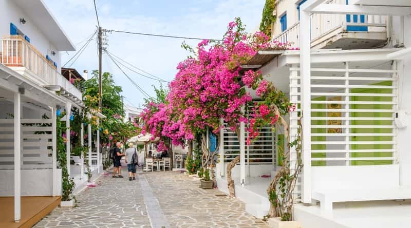 A photograph capturing a street scene in Antiparos, Greece, featuring traditional Greek architecture typical of the Cyclades islands. The image showcases the characteristic white-washed buildings with blue accents against a clear sky. 