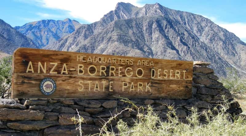 A photograph depicting a wooden stele with the inscription 'Anza-Borrego Desert State Park' affixed to stonework. The image is situated against the backdrop of a mountain. The stele serves as a marker, providing information about the location and denoting the boundary of Anza-Borrego Desert State Park.