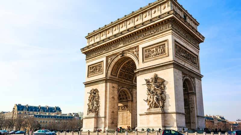 The image captures a sunny view of the Arc de Triomphe in the Étoile Square in Paris, France. The iconic monument stands prominently in the center of the square. Surrounding the Arc de Triomphe are wide avenues and bustling city streets.