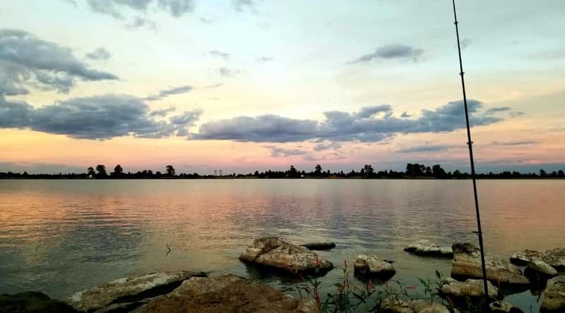 A photograph depicting Ashurst Lake in Arizona. The image captures the lake's landscape with clear waters. The shoreline features greenery and is framed by a backdrop of trees.