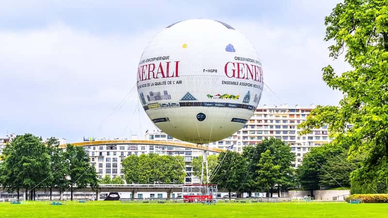 Parc André Citroën in Paris, France, with a tethered hot air balloon named "Air de Paris." The balloon is notably large and white, adorned with inscriptions. It is tethered securely to the ground, providing a static focal point in the park. Surrounding the balloon are extensive greenery and landscaping.