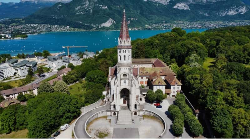 Aerial view of the Basilique de la Visitation surrounded by a lake, picturesque Alps and green lush trees. This elegant church stands out with its pristine white façade, ornate detailing, and a graceful dome that gleams in the sunlight.