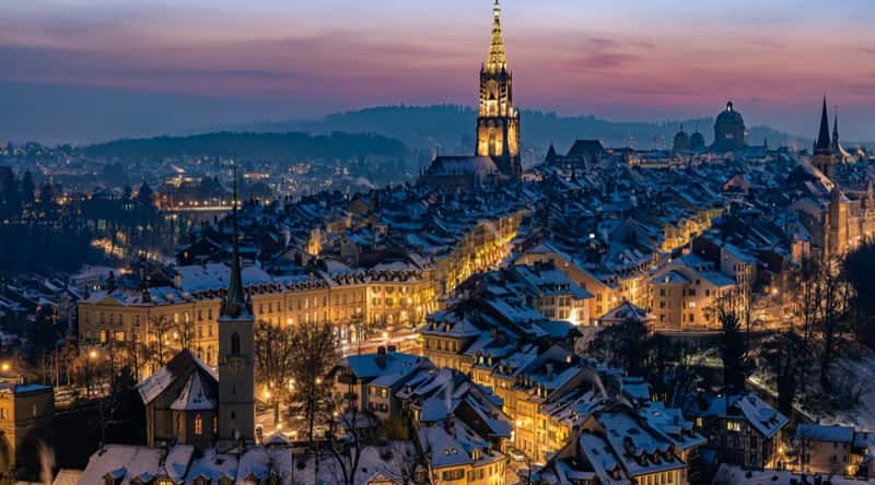 An aerial view capturing the nocturnal cityscape of Bern, Switzerland. The image displays streets illuminated by yellow lanterns, creating a warm glow against the dark backdrop. The roofs of houses are covered with snow. 
