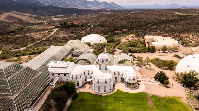 The image depicts the exterior of the University of Arizona Science campus, specifically highlighting Biosphere 2 in Oracle, Arizona. The structure appears modern and sleek, with a combination of glass panels and metallic elements. The color palette consists of neutral tones, blending harmoniously with the desert surroundings typical of Arizona.