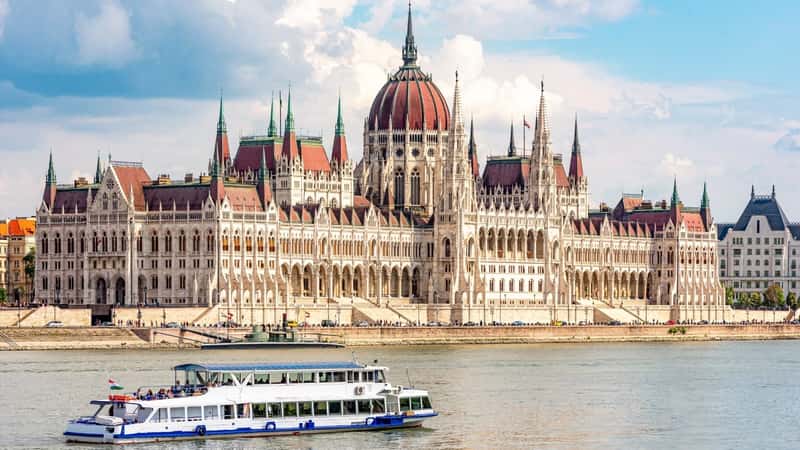 A photograph capturing the Hungarian Parliament Building situated along the banks of the Danube River, with a passing excursion boat in view. The image illustrates the iconic architecture of the parliament building against the backdrop of the river.