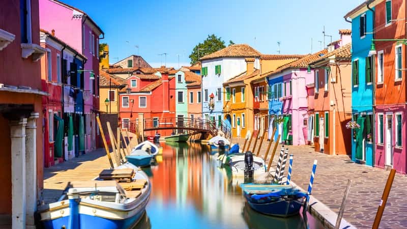 The image portrays a scene of colorful houses and buildings along the riverbank, their vibrant hues creating a picturesque view. The reflection of these buildings in the river appears blurry, adding a sense of movement and fluidity to the scene.