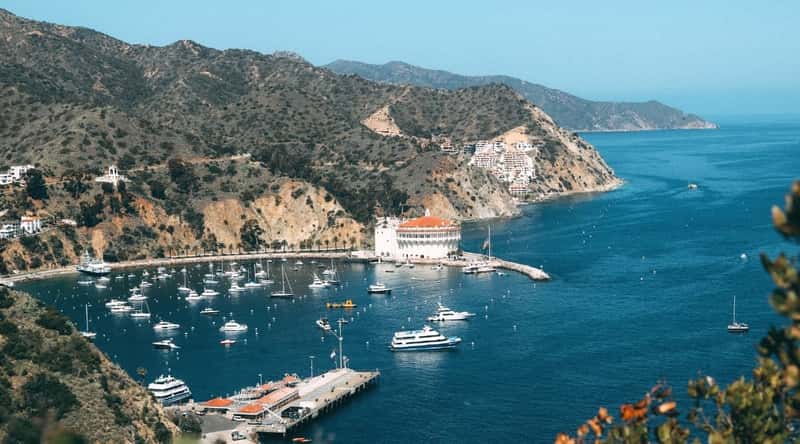 An aerial photograph capturing Catalina Island's topography, characterized by numerous mountains and hills. The image features a pier with an array of boats and ships docked.