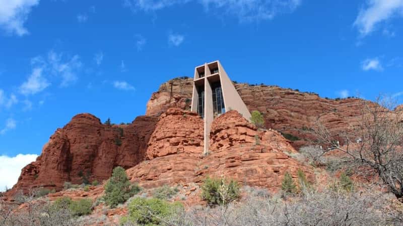 A photograph capturing the Chapel of the Holy Cross in Sedona, Arizona. The image showcases the chapel's distinctive architecture against the backdrop of the surrounding red rock formations. The chapel appears as a focal point in the landscape, with minimal vegetation and clear skies.