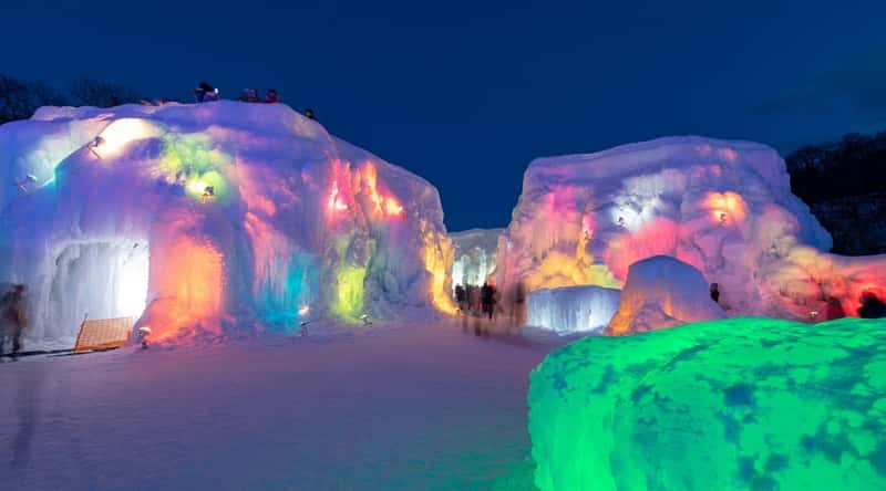 A photograph capturing scenes from the Chitose - Lake Shikotsu Ice Festival. The image showcases various ice sculptures and structures against the backdrop of the festival grounds. Visitors, both locals and tourists, can be seen observing the frozen artworks.