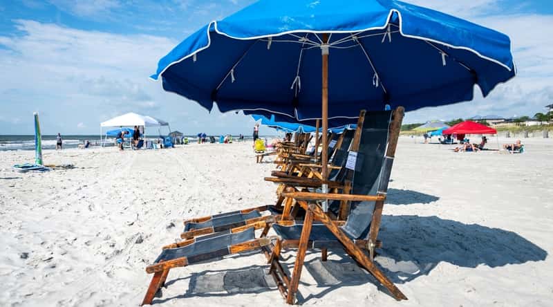 Coligny Beach Park is a public beach park located in Hilton Head Island, South Carolina. It features a beautiful sandy beach, clean waters, and a wide range of amenities. Close-up of wooden sun loungers with blue umbrellas on a white sand beach.