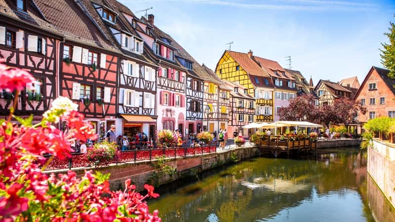 The image showcases a water canal in Dijon, France, bordered by traditional half-timbered houses. The houses are characterized by their cleanliness, brightness, and vibrant colors, adding to the visual appeal of the scene.
