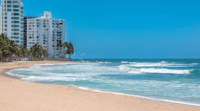 A photograph capturing Condado Beach in Puerto Rico. The image features a stretch of sandy shoreline. The turquoise waters of the Atlantic Ocean gently meet the sandy expanse. High-rise buildings form the backdrop, showcasing the urban setting around Condado Beach.
