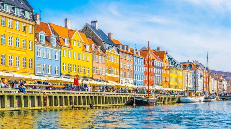The image presents a view of the old Nyhavn port in central Copenhagen, Denmark. The houses along the port are distinctly colored in a row, featuring shades of yellow, orange, blue, and red, creating a vibrant and picturesque waterfront scene. The area is bustling with many people walking along the alley in front of the houses, enjoying the waterfront ambiance.