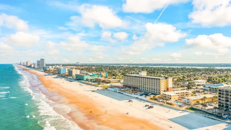 Drone photos of Daytona Beach, Florida, reveal beautiful beaches with striking blue water. The ocean lies to the left, with its deep, vibrant hues contrasting against the sandy shore. On the right, the beach extends into the city, where buildings and streets are visible. The beach itself is wide and sandy, with a mix of beachgoers and umbrellas dotting the shoreline.