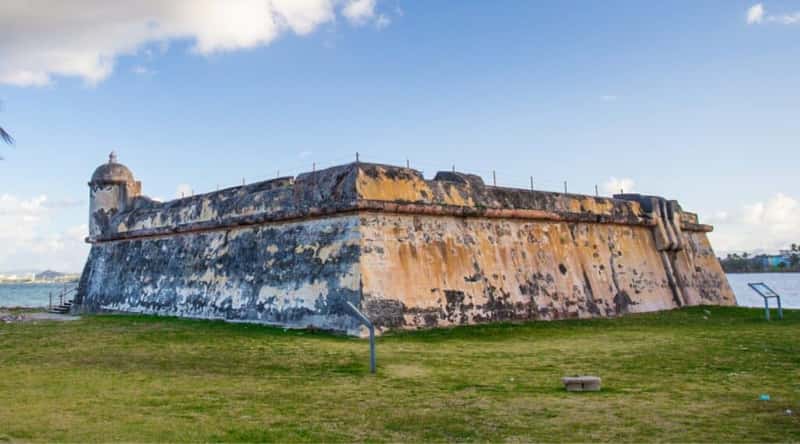 A photograph capturing Fortín San Juan de la Cruz in Puerto Rico. The image features the historic fortress situated against a coastal backdrop. The fort exhibits traditional military architecture with solid stone walls and structures. 