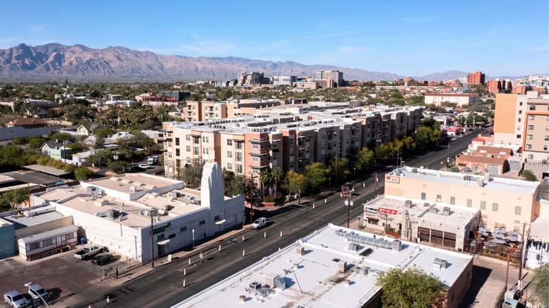 The image depicts Tucson, Arizona, USA, specifically focusing on the Historic Fourth Avenue neighborhood. In the scene, the afternoon sun illuminates the neighborhood, casting a warm glow on the buildings and streets. The architecture consists of charming, low-rise structures.