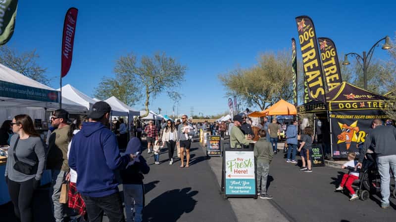 A photograph capturing the scene at Gilbert Farmers Market, depicting numerous people walking along the counters.