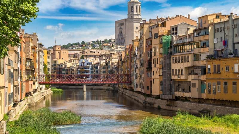 The image features colorful houses alongside the Eiffel bridge, with the Saint Mary Cathedral in the background, capturing a beautiful summer day in Girona, Catalonia, Spain. The vibrant hues of the houses add a lively contrast to the scene.