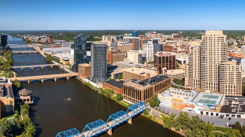 In Grand Rapids, Michigan, the city view includes a wide river crossed by six bridges in the foreground. The cityscape features tall buildings. Beyond the city, there is a significant amount of greenery visible, suggesting natural surroundings outside the urban center.