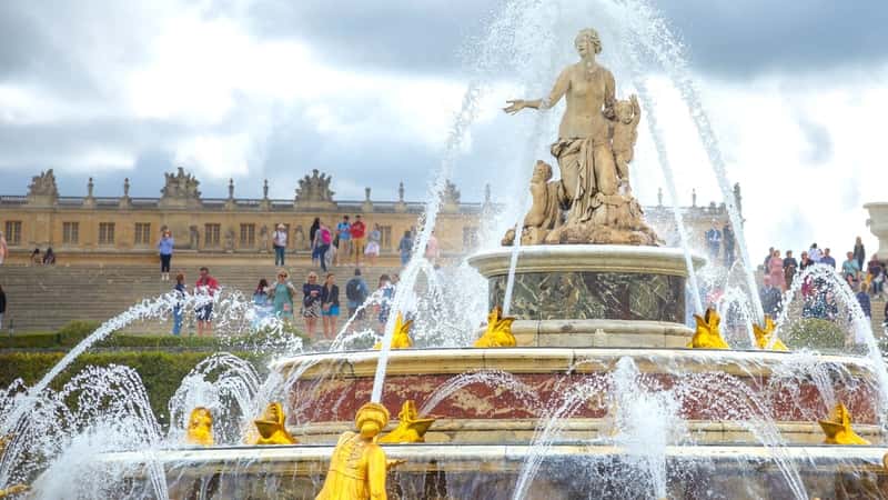 The image shows the Latona Fountain in Paris, France, with the Versailles Palace and tourists in the background. The fountain is a focal point, featuring classical sculptures and water features. The palace's grand architecture is visible behind the fountain.