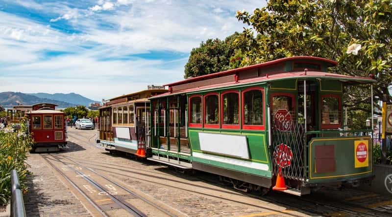Green trolley car riding along the railway in San Francisco. The train is green, with red cut outs around the windows. It is in a stopped position, waiting for its next route.