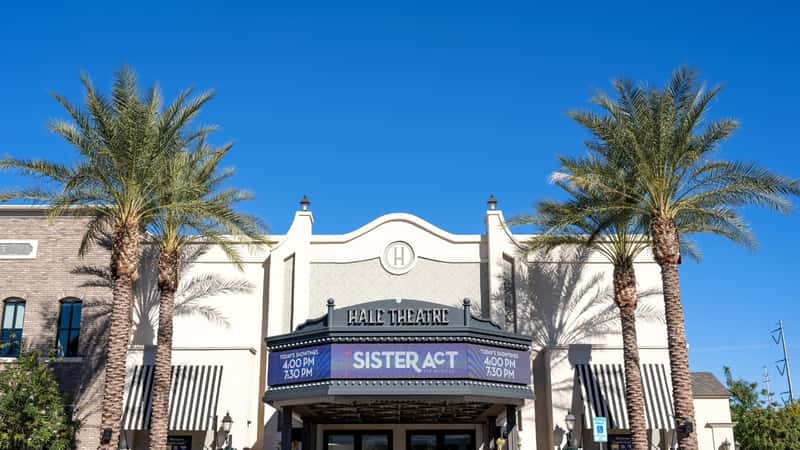 A view capturing Hale Center Theater in Gilbert, Arizona. The image shows a sign reading 'Hale Theatre' in the foreground, with four palm trees nearby. The sky is clear, and the building, light in color, appears almost white. The architectural design of the theater is visibles.