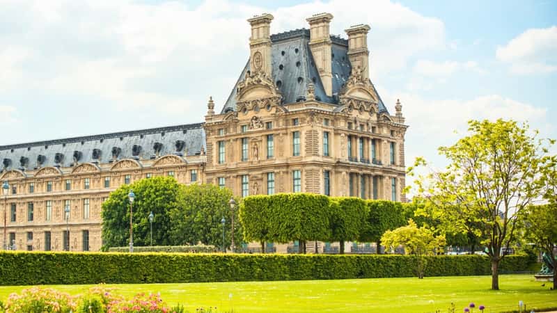 Jardin des Tuileries in Paris, France. The foreground features well-groomed lawns, meticulously trimmed bushes and trees, and squared-off shrubbery. In the background, there is a prominent large historical building, adding a sense of grandeur and historical significance to the scene.