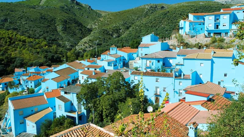 The image showcases Juzcar, Spain, known as the "blue village," set against a backdrop of green mountains. The village's distinctive feature is its blue-painted houses with brown roofs, creating a unique and colorful visual contrast against the natural landscape. The mountains surrounding the village are lush and verdant.