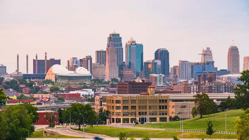 The Kansas City skyline at sunset features a blend of modern and historic buildings. The cityscape includes tall skyscrapers with reflective glass exteriors and shorter, older structures with brick facades. The sunset casts a warm, golden hue over the buildings, enhancing the visual appeal.