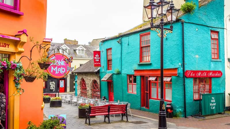 The image depicts Market Street and Newman's Mall in Kinsale, Cork, Ireland, showcasing a row of colorful houses. Among them are houses painted in vibrant hues, such as orange with pink windows and another in green with red windows and doors.