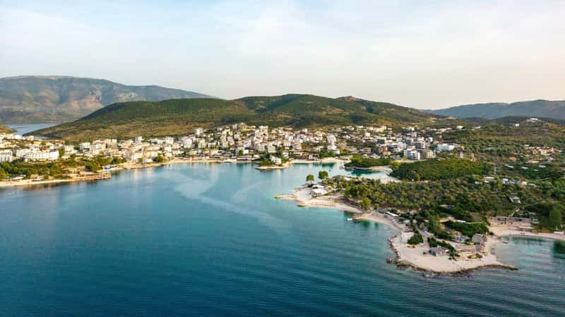 An aerial photograph capturing the Bay of Ksamil in the Albanian Riviera. The image showcases the coastal landscape featuring clear blue waters and a shoreline with identifiable geographic formations. The bay is surrounded by hills and greenery.